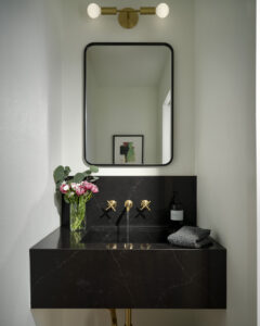 Modern bathroom with a black marble sink and gold fixtures. A rectangular mirror hangs above, with a vase of pink flowers and a candle on the counter. Soft lighting and a small framed picture on the wall create a stylish ambiance.